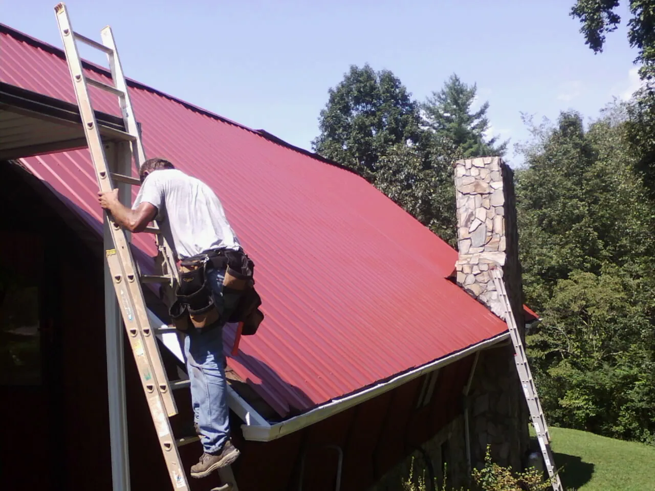 Roofer on a ladder beside a red metal roof and stone chimney — Soffit & Fascia Repair by Roofing Brothers in Pueblo